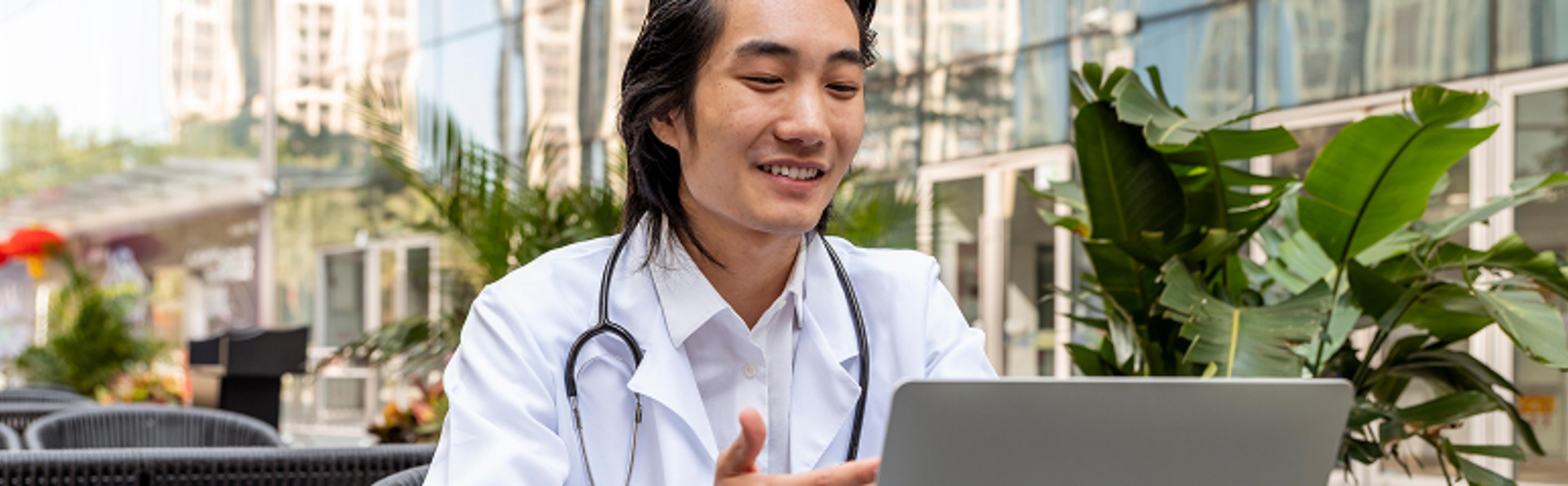 Individual in white coat with stethoscope using laptop outdoors near plants and buildings