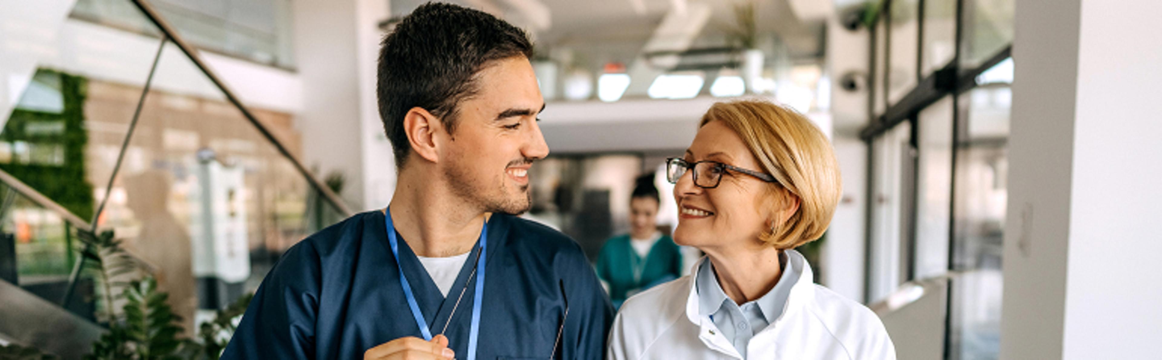Two medical professionals walking through hospital corridor, one in blue scrubs, one in white coat