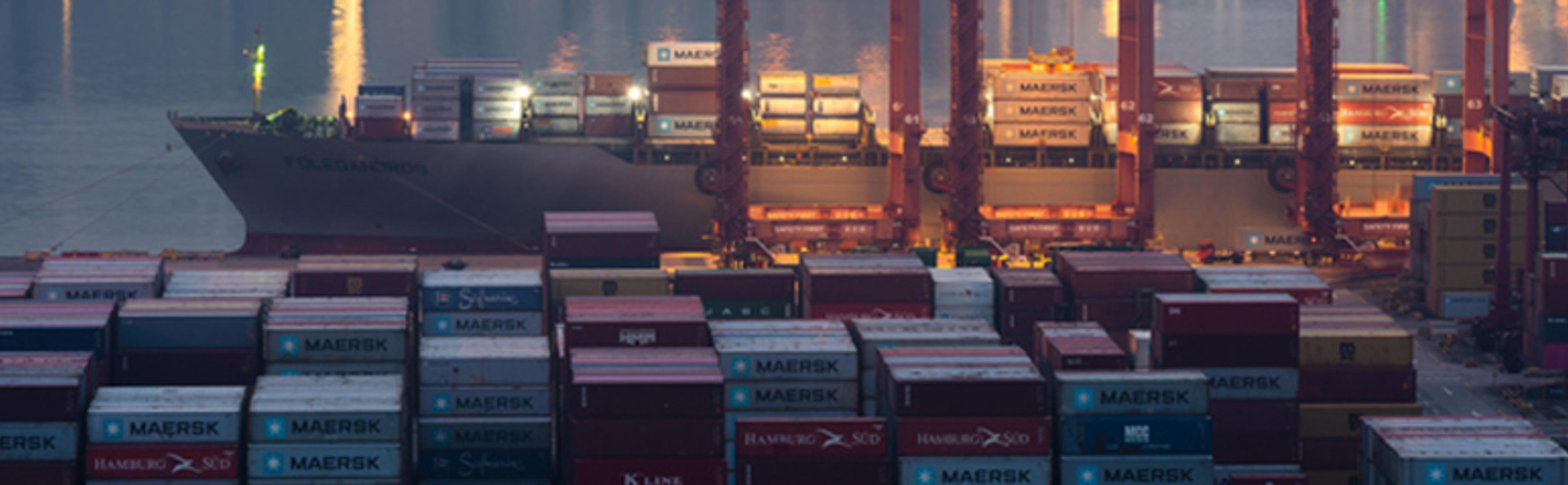 A low-angle photo of a container ship docked at a large, brightly lit port at dusk