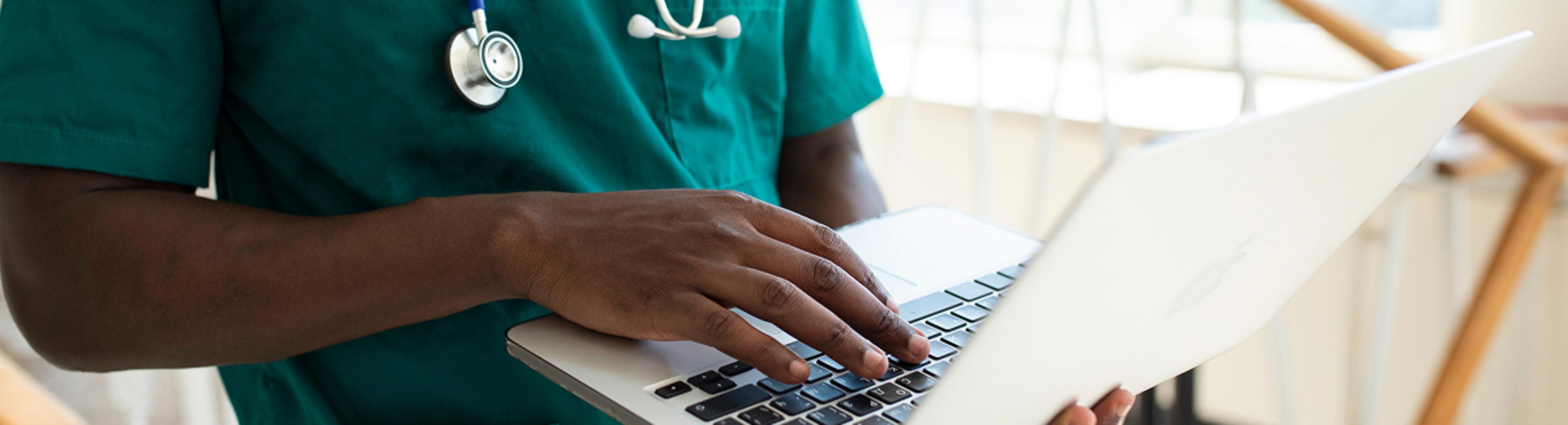 A medical professional wearing green scrubs with a stethoscope types on a laptop computer