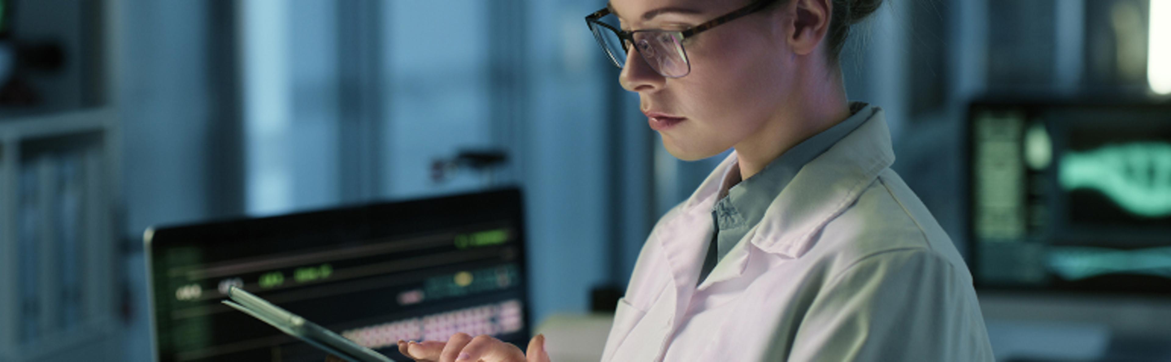 A person wearing a white lab coat and glasses is holding a tablet in a laboratory setting. In the background, computer screens display various data and images, including medical imaging on one of the monitors