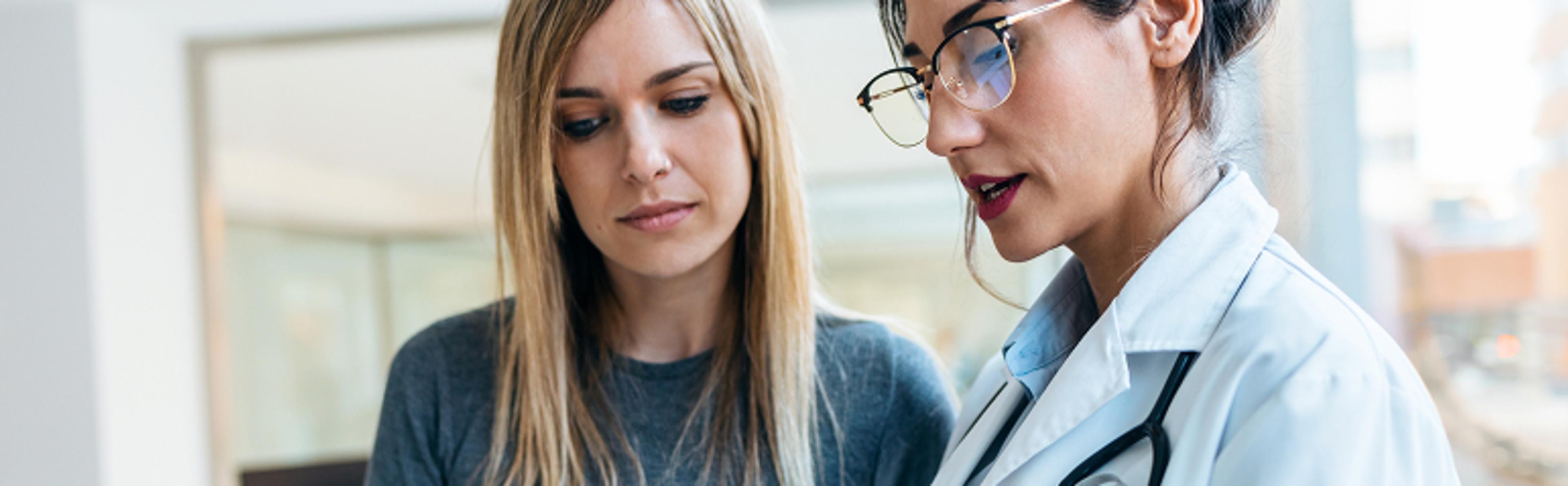 Doctor with stethoscope talking to person with long hair in gray shirt indoors