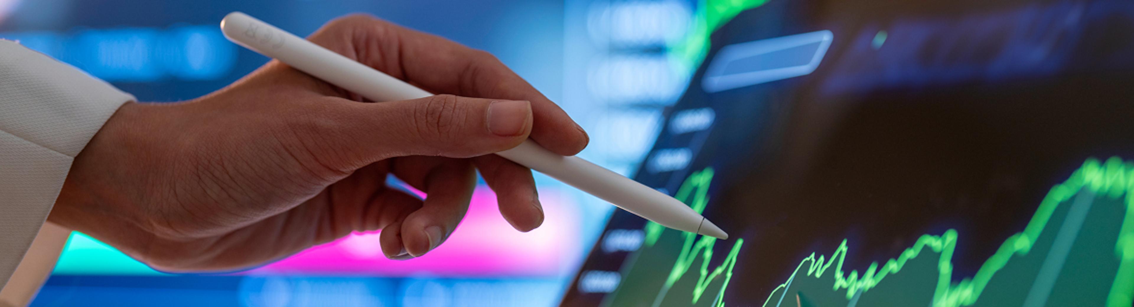 Hand with stylus pointing at digital screen showing green line graphs and charts