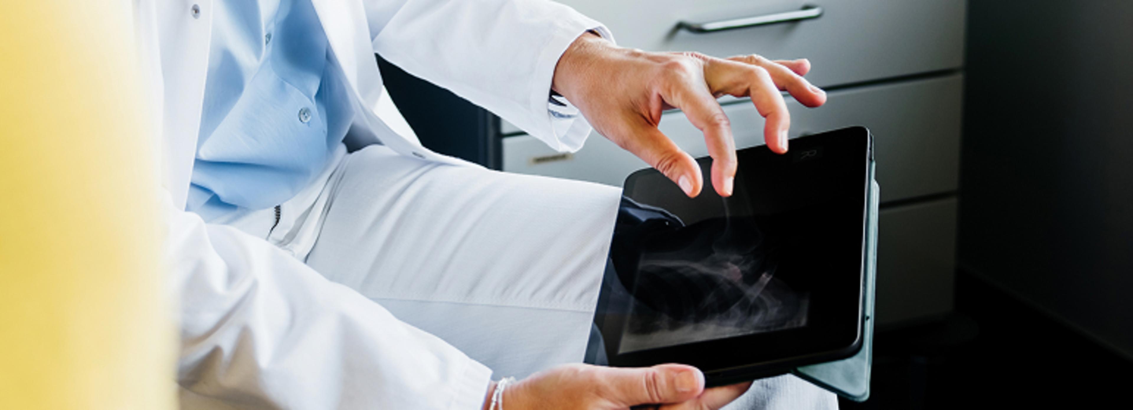 A person in a white lab coat is holding a tablet with both hands, using the right hand to interact with an X-ray image displayed on the screen. The person's left leg is visible, and drawers are seen in the background