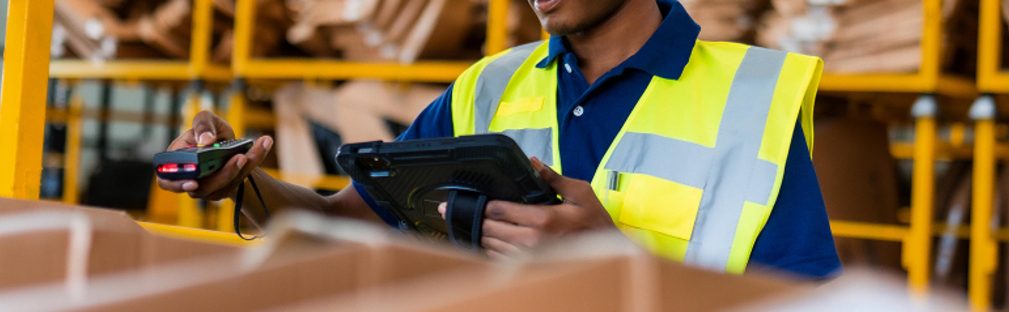 A warehouse worker wearing a high-visibility yellow vest is scanning a barcode on a package with a handheld scanner while holding a tablet. Shelves filled with cardboard boxes and packages are visible in the background