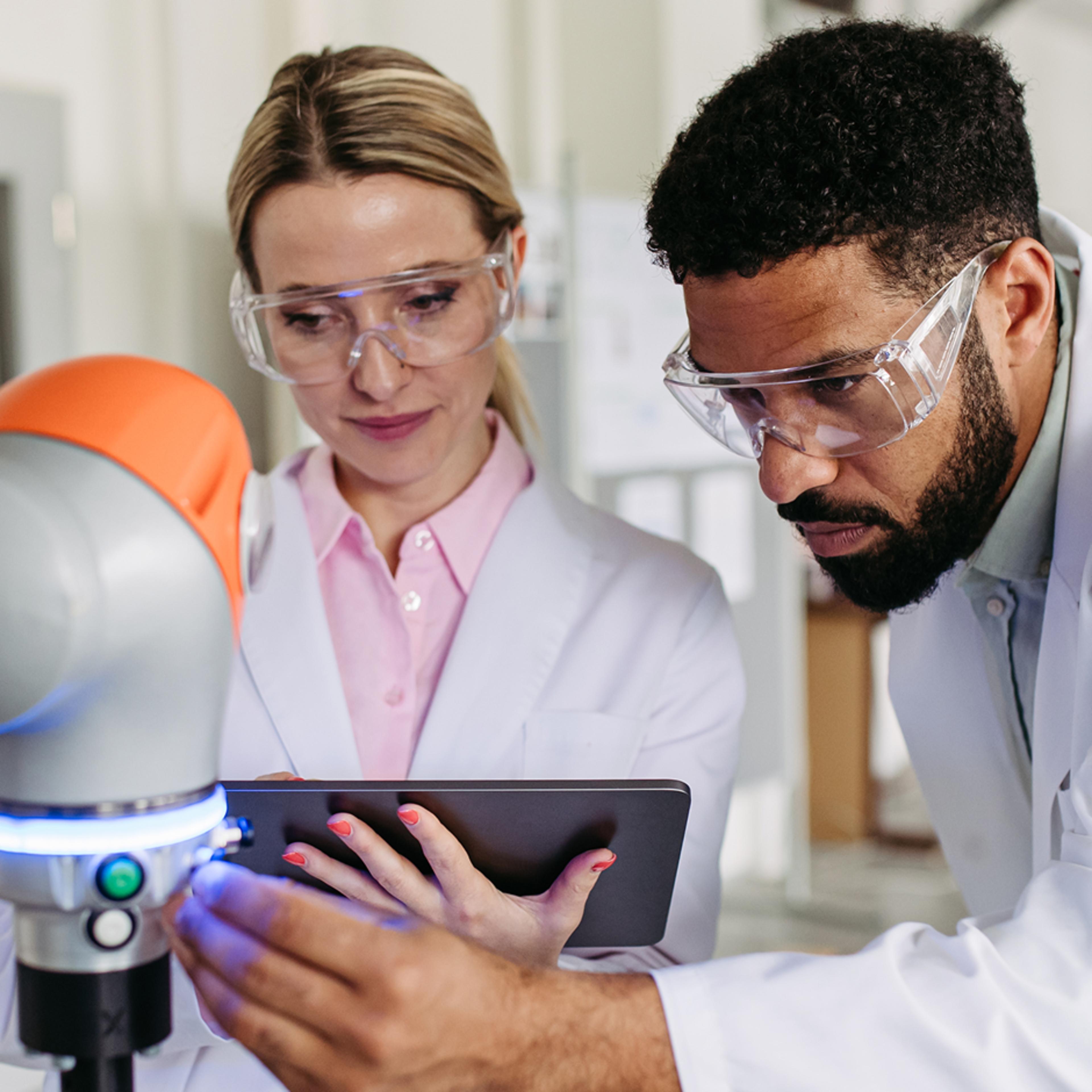 Two medical professionals, both wearing safety glasses and lab coats, closely examine a robotic arm in a collaborative, high-tech laboratory environment
