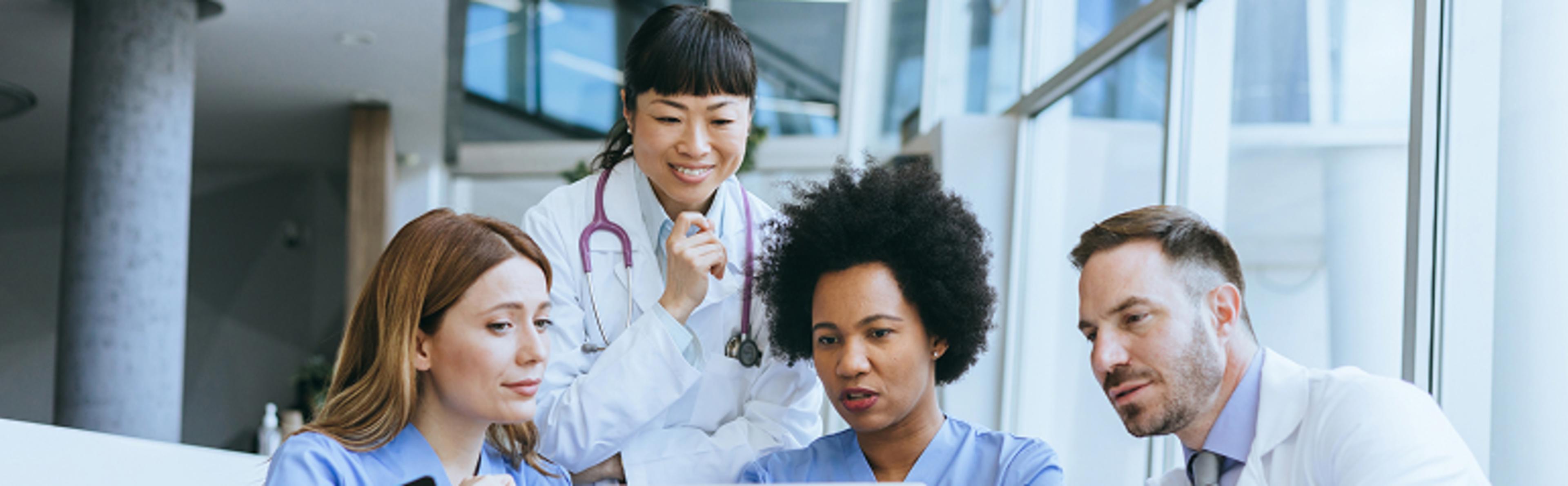 Four medical professionals in white coats and blue scrubs having a discussion with faces obscured