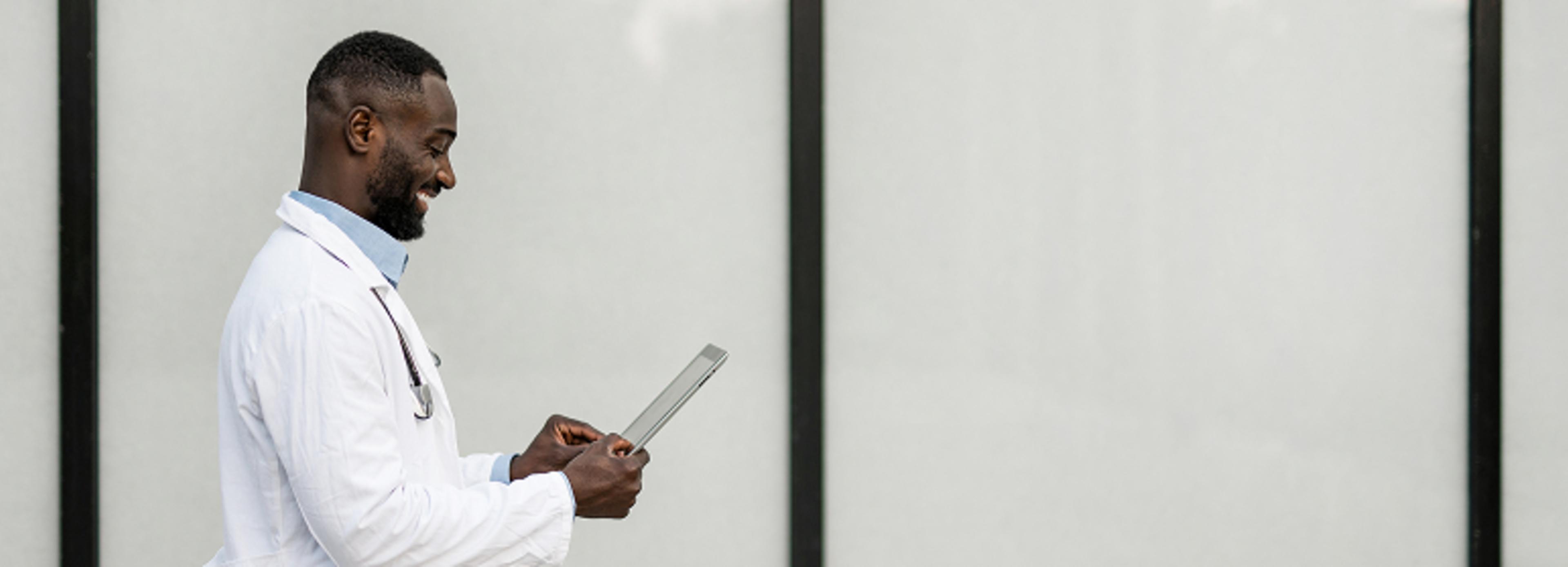 A person in a white lab coat with a stethoscope around the neck is holding and looking at a tablet. The background features large, light-colored panels with dark vertical lines