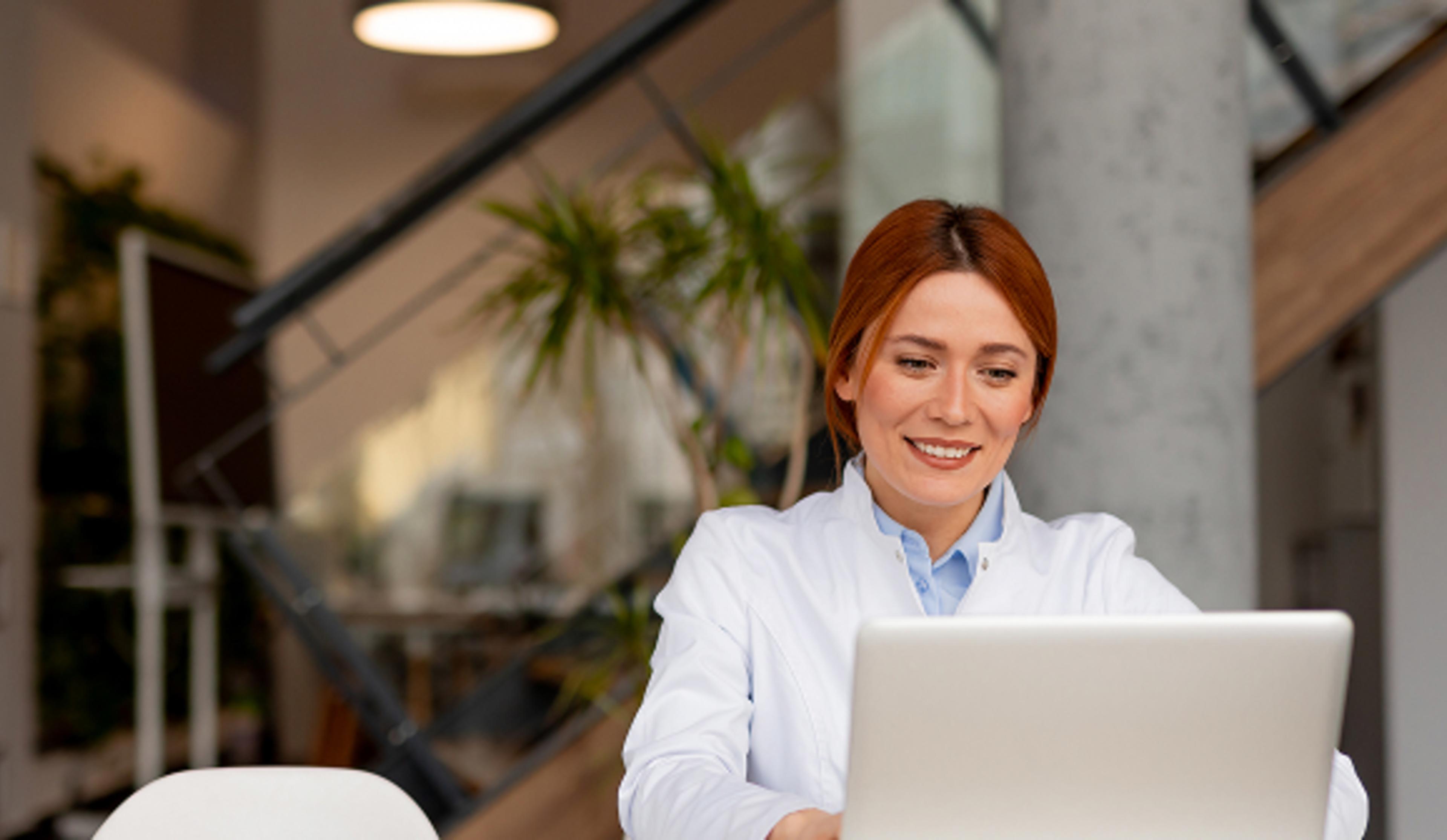 Individual in white coat working on laptop in modern office with plants and stairs in background