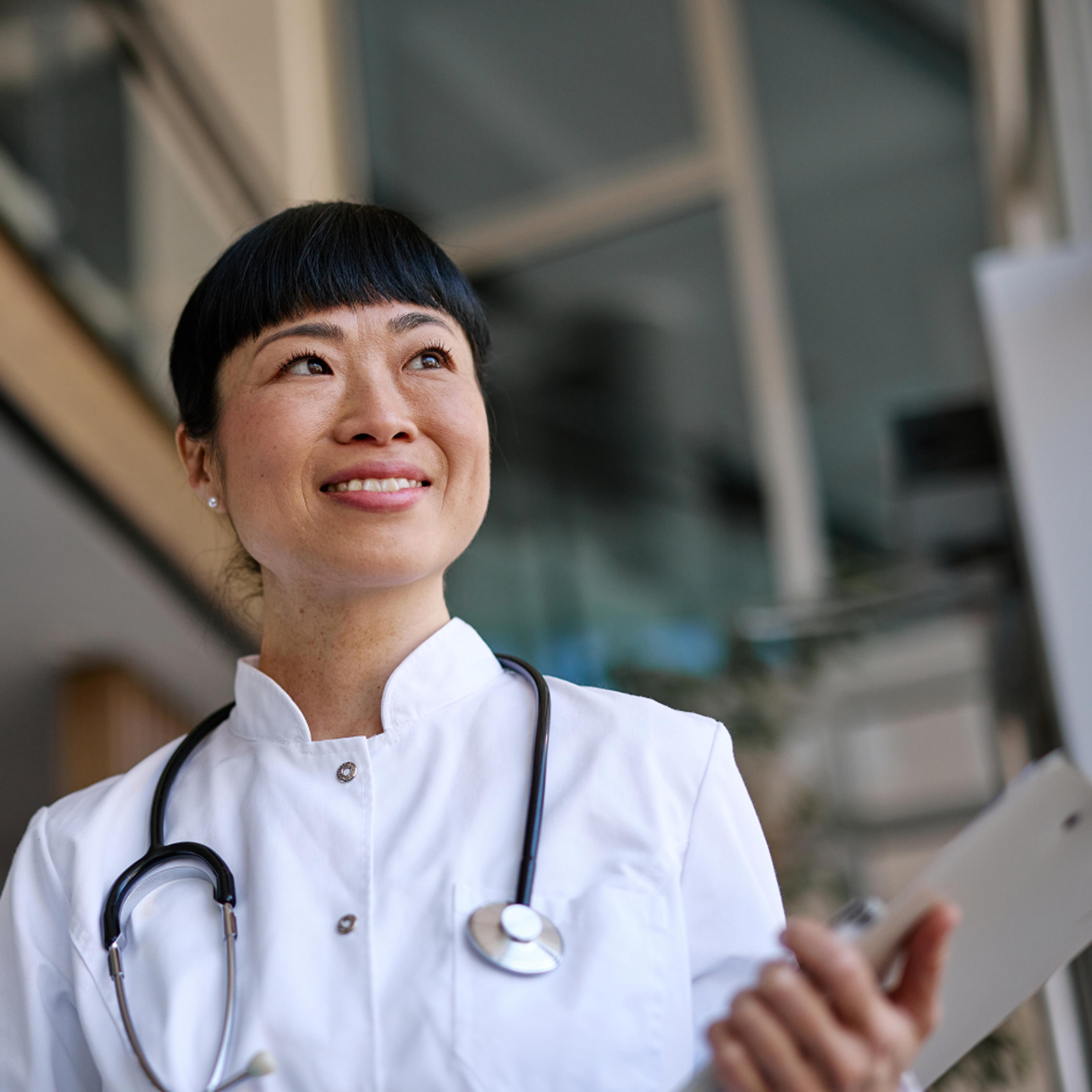 A medical professional with a stethoscope and a clipboard smiles and looks into the distance