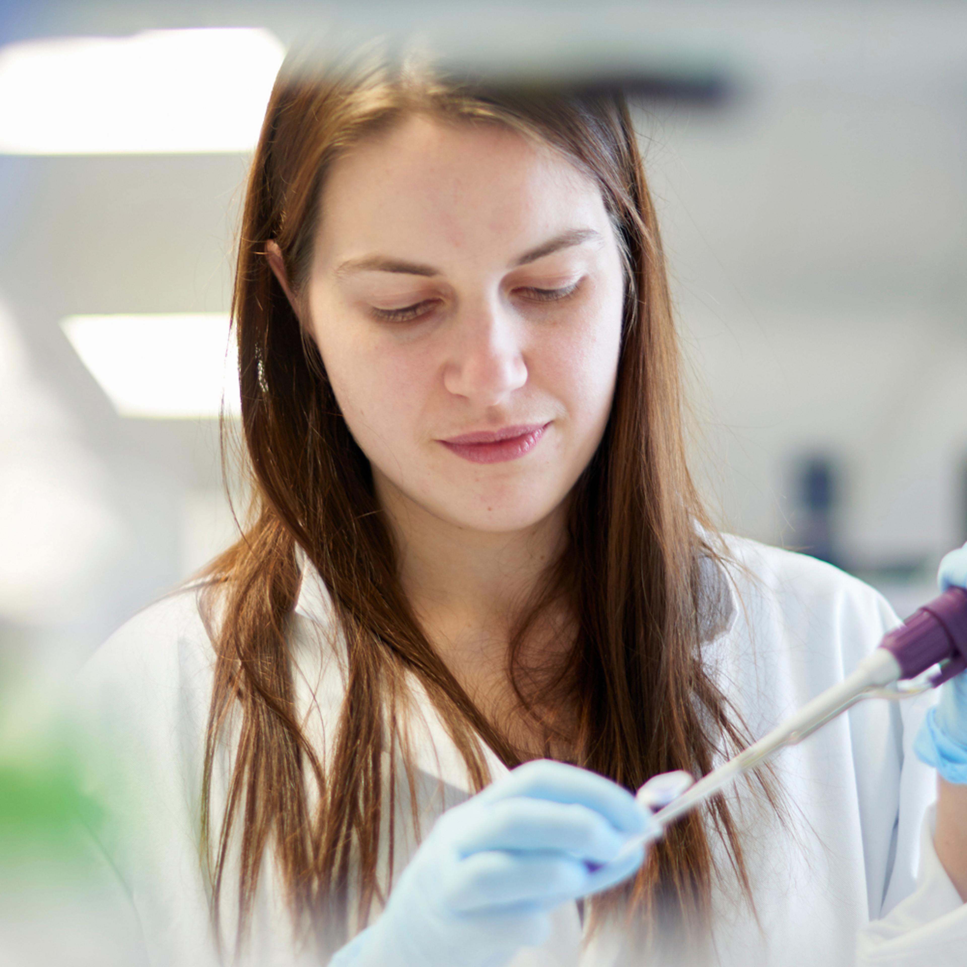 A medical professional in a white lab coat carefully pipetting a blue liquid into a small vial in a bright, modern laboratory