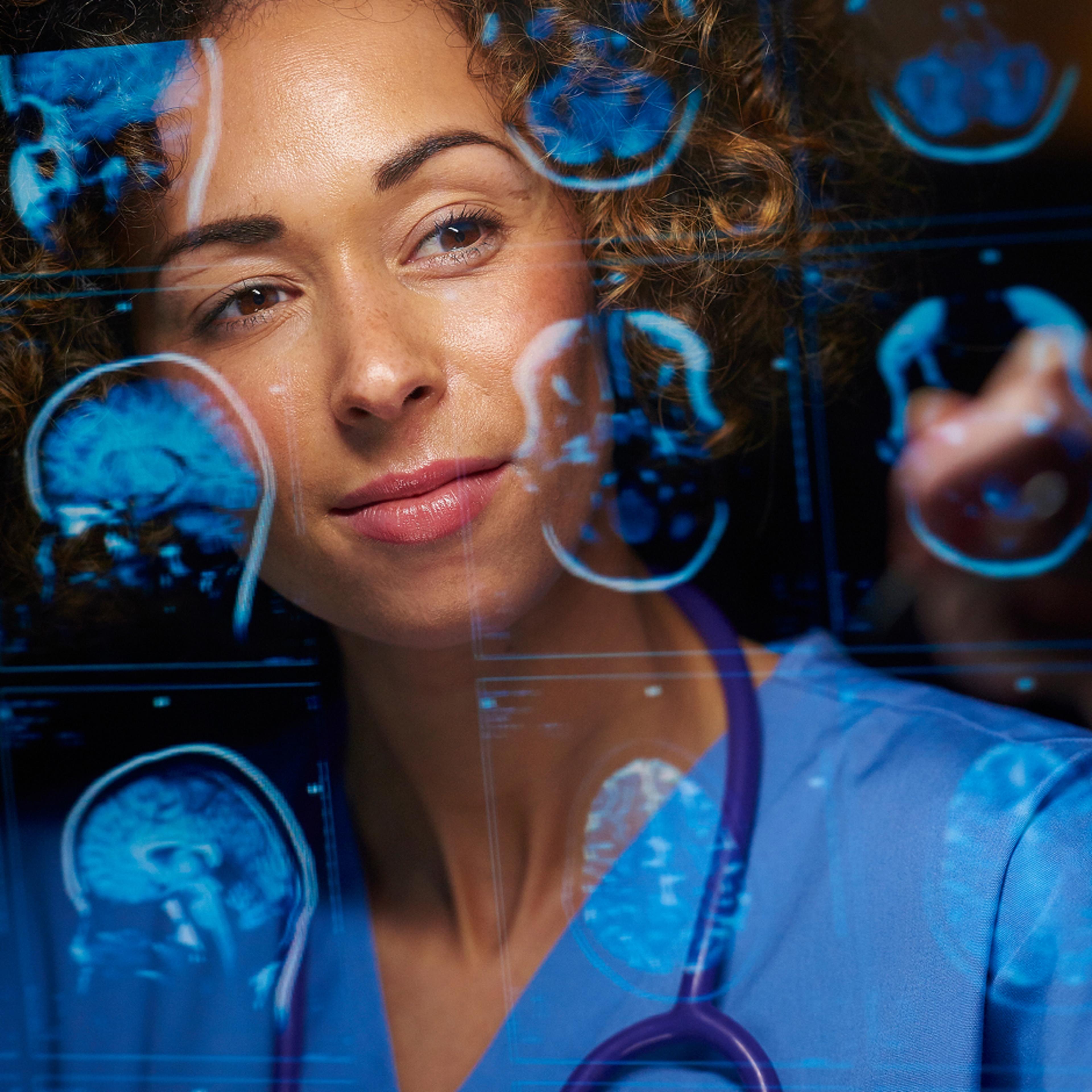 A medical professional wearing a blue scrub top and a stethoscope, looking at a transparent screen that displays multiple blue-toned medical scans and data