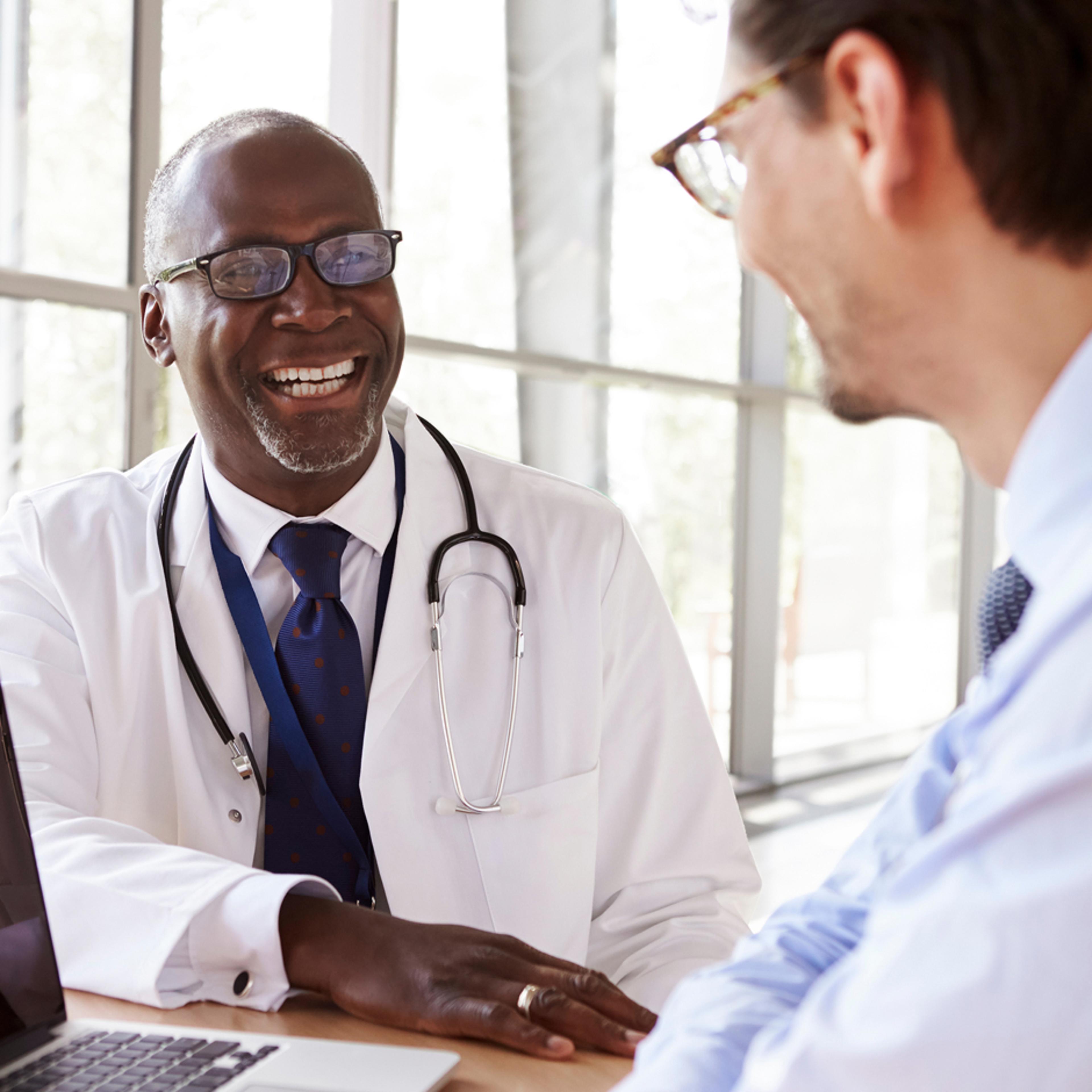 A smiling doctor in white lab coat, wearing glasses and stethoscope. He is at a desk with a laptop and engaged in friendly conversation with a patient