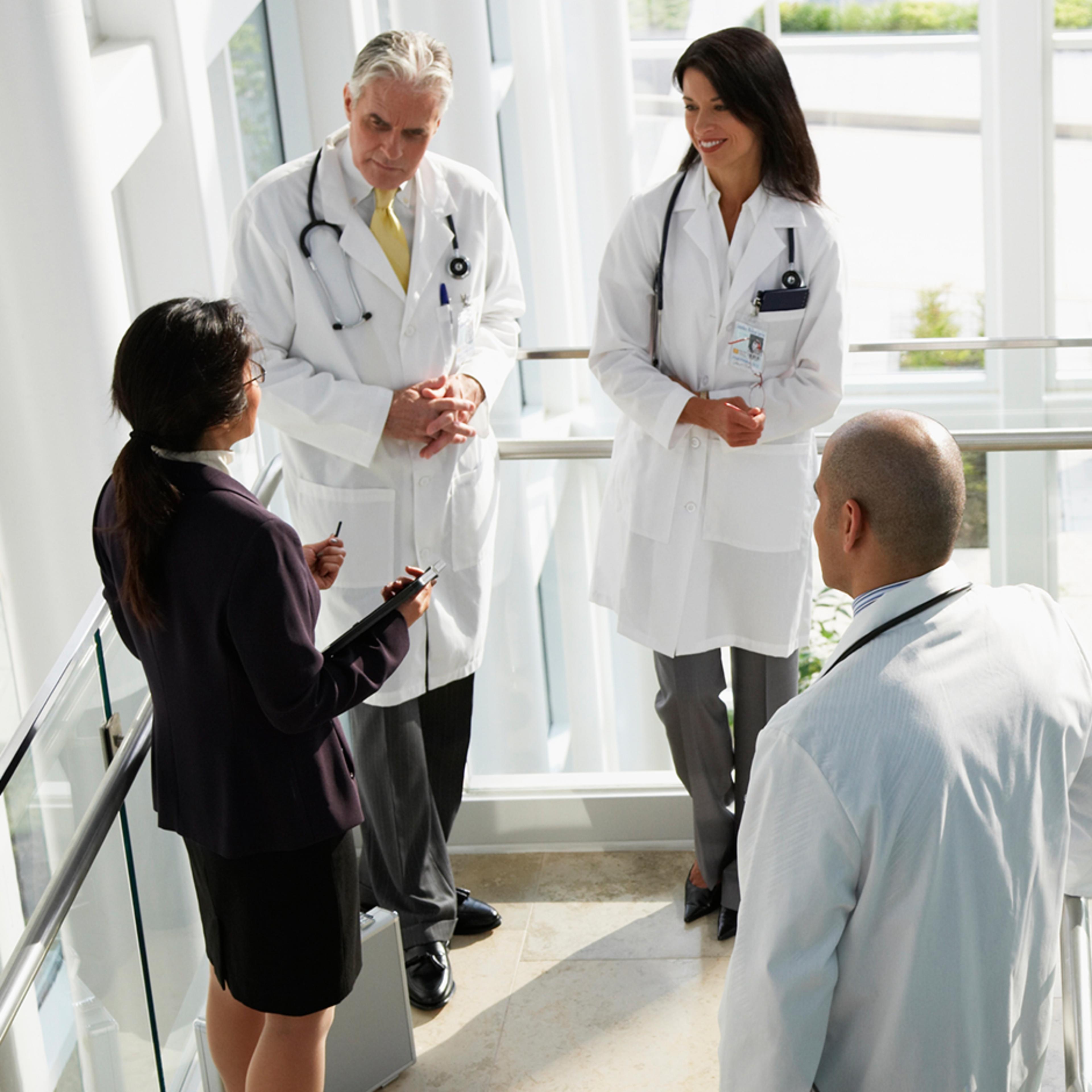 Three healthcare professionals, wearing white lab coats and stethoscopes, and an executive standing in a modern, well-lit hospital, engaged in a discussion