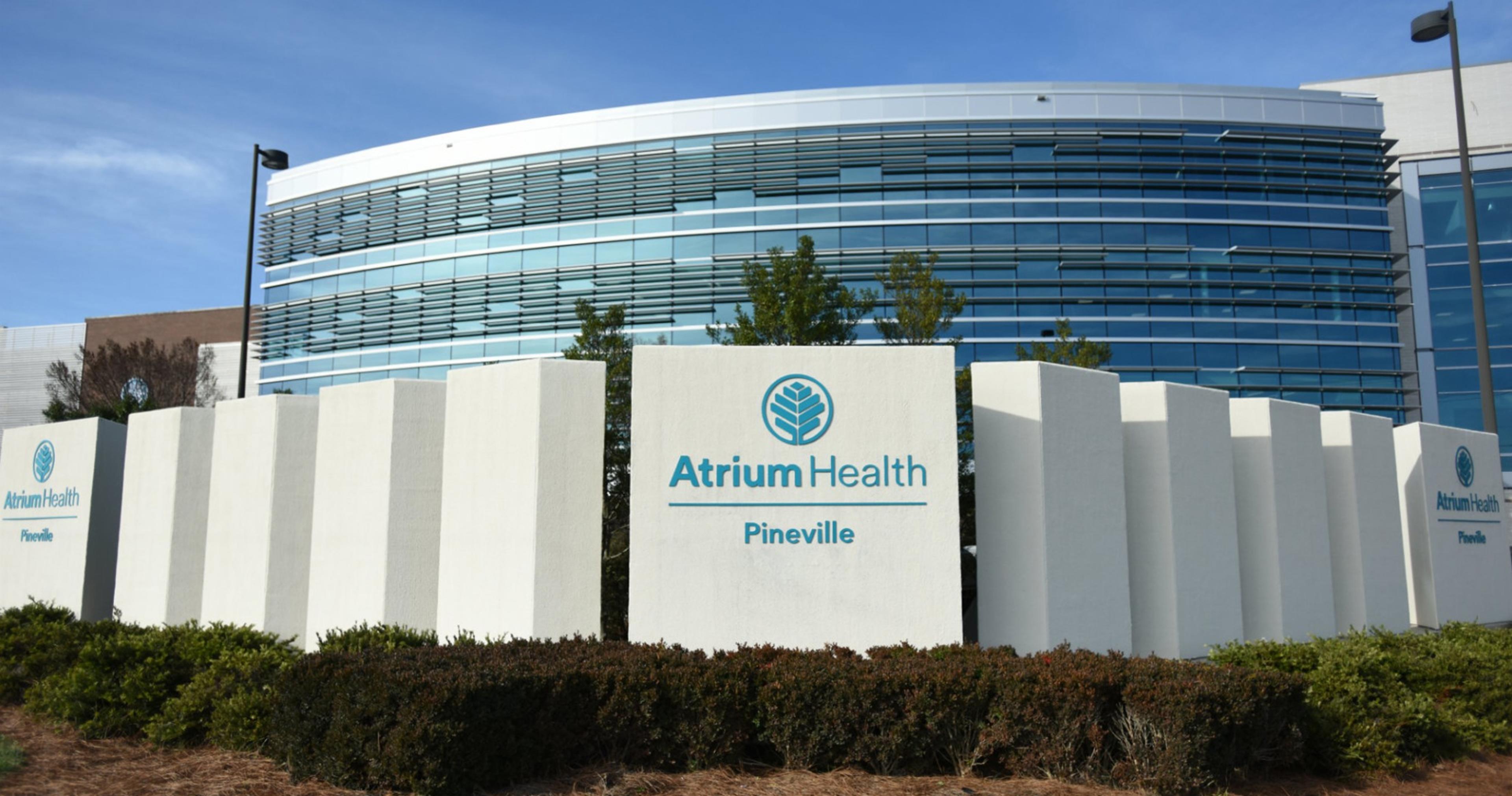 The exterior of Atrium Health Pineville, a healthcare facility. The building has a modern design with large glass windows and a curved structure. In front of the building, there is a sign with the Atrium Health logo and the text 'Atrium Health Pineville' displayed prominently. There are also some bushes and greenery in front of the sign