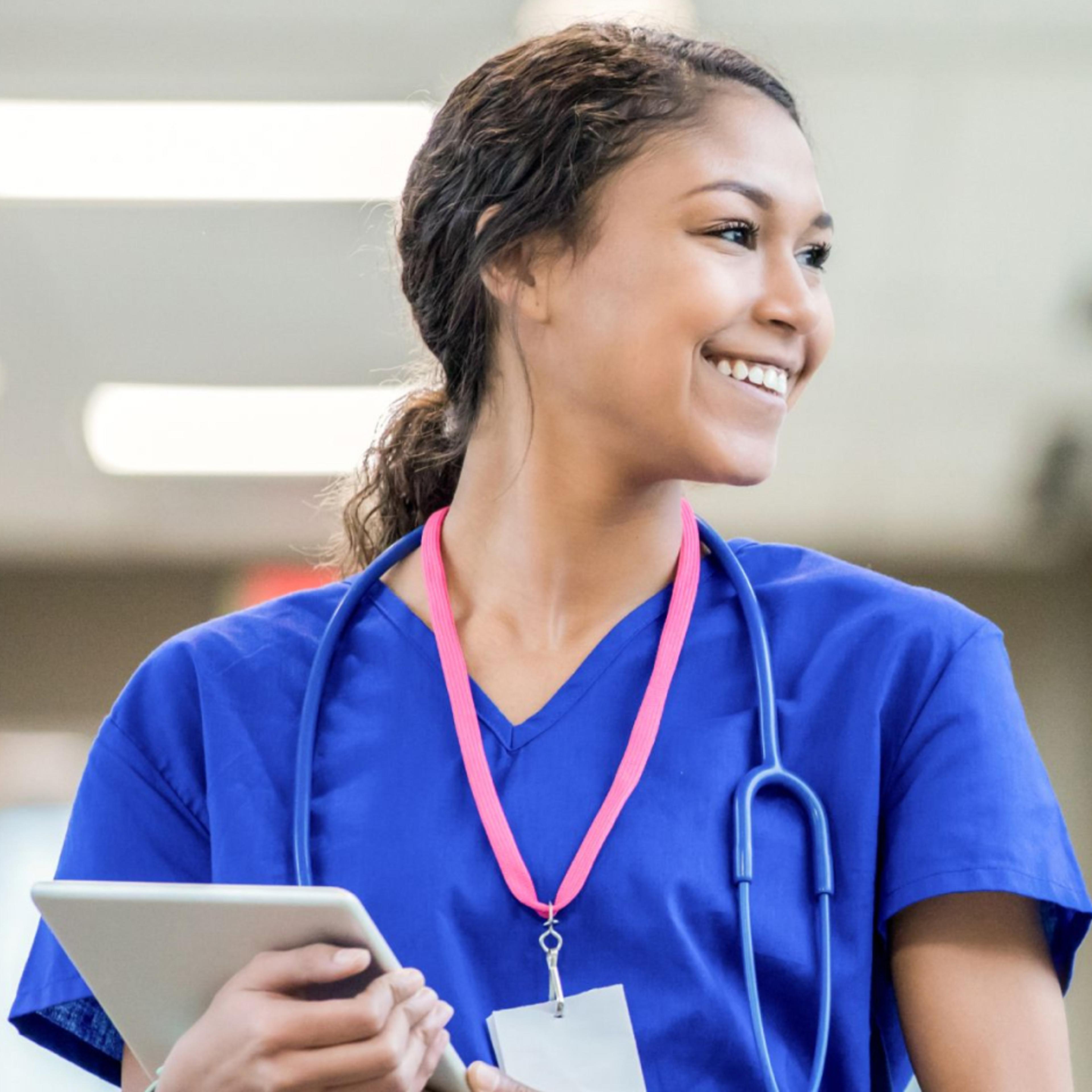 Person in blue scrubs with stethoscope holds a tablet and looks to the side