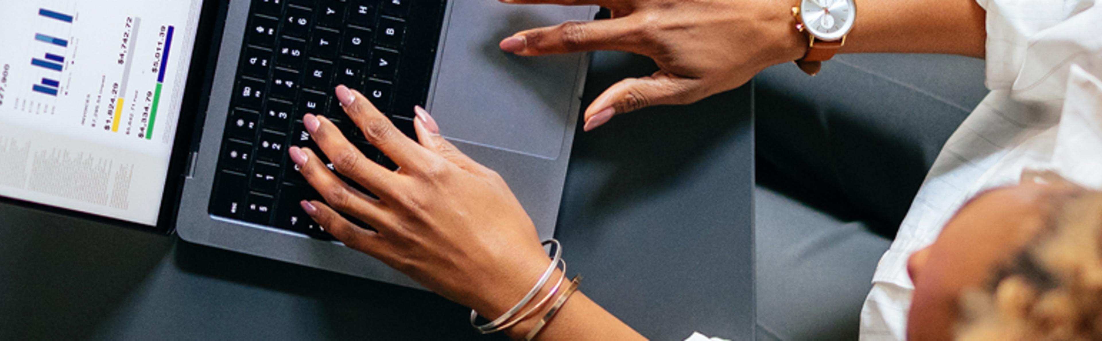 Hands typing on laptop keyboard with document showing charts and graphs nearby