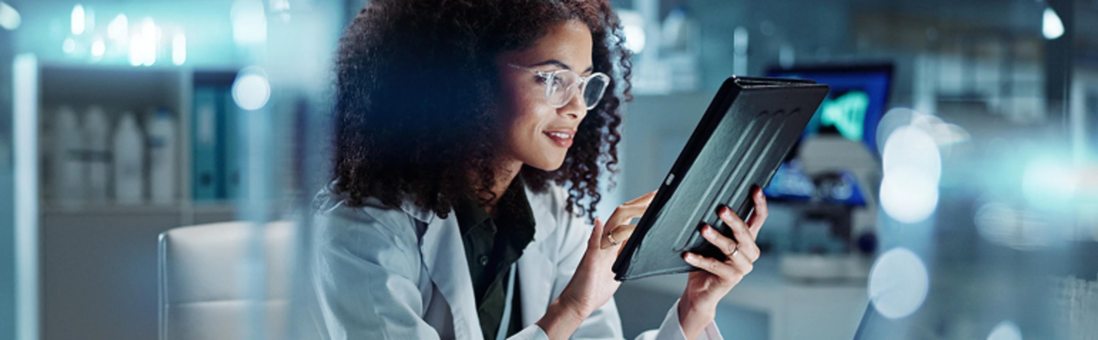 A person in a white lab coat is sitting in a laboratory setting, holding and examining a tablet device. The background is blurred, showing various lab equipment and shelves