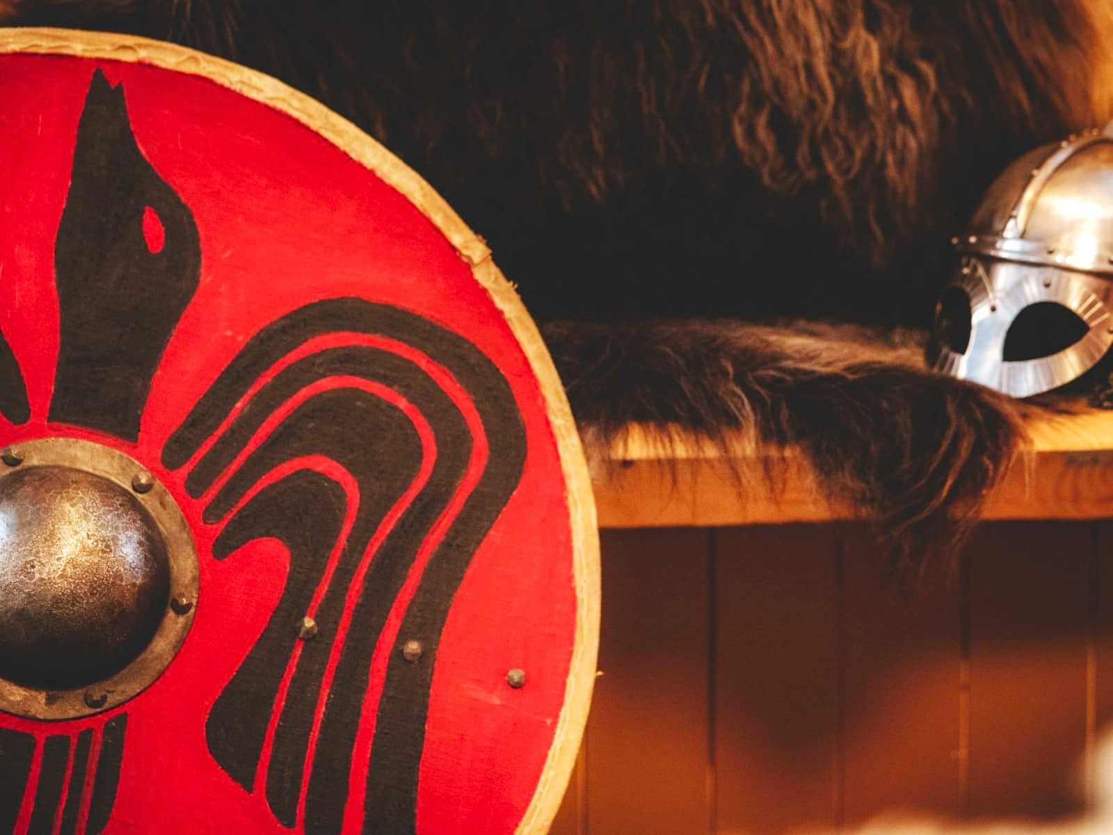 Red Viking shield with black ornaments and Viking helmet in the background.