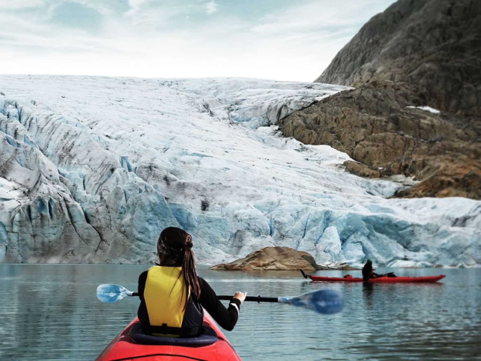 Eine Person in einem roten Kajak, von hinten gesehen, paddelt vor einem Gletscher mit einem anderen Kajak im Hintergrund.