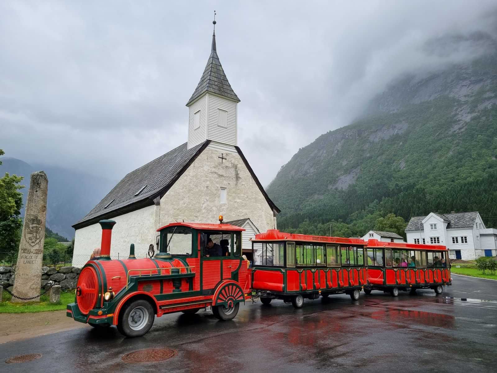 Red and green mini train on wheels (the Trolltrain) parked in front of Eidfjord church.