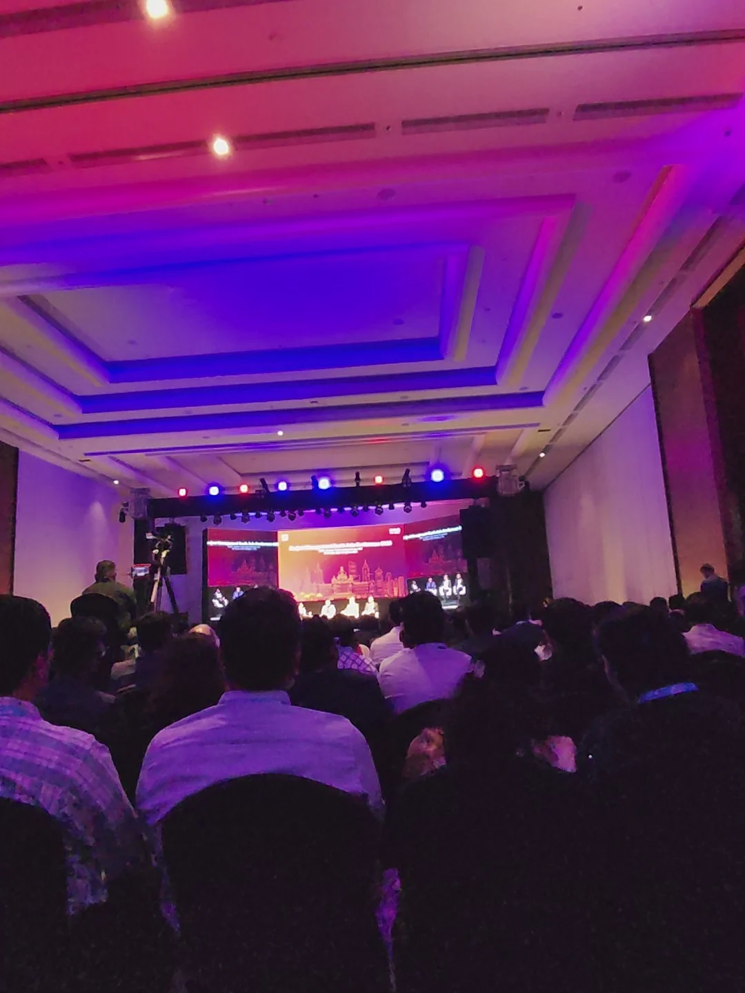 A large audience sits facing a brightly lit stage with a wide screen at an indoor event.
