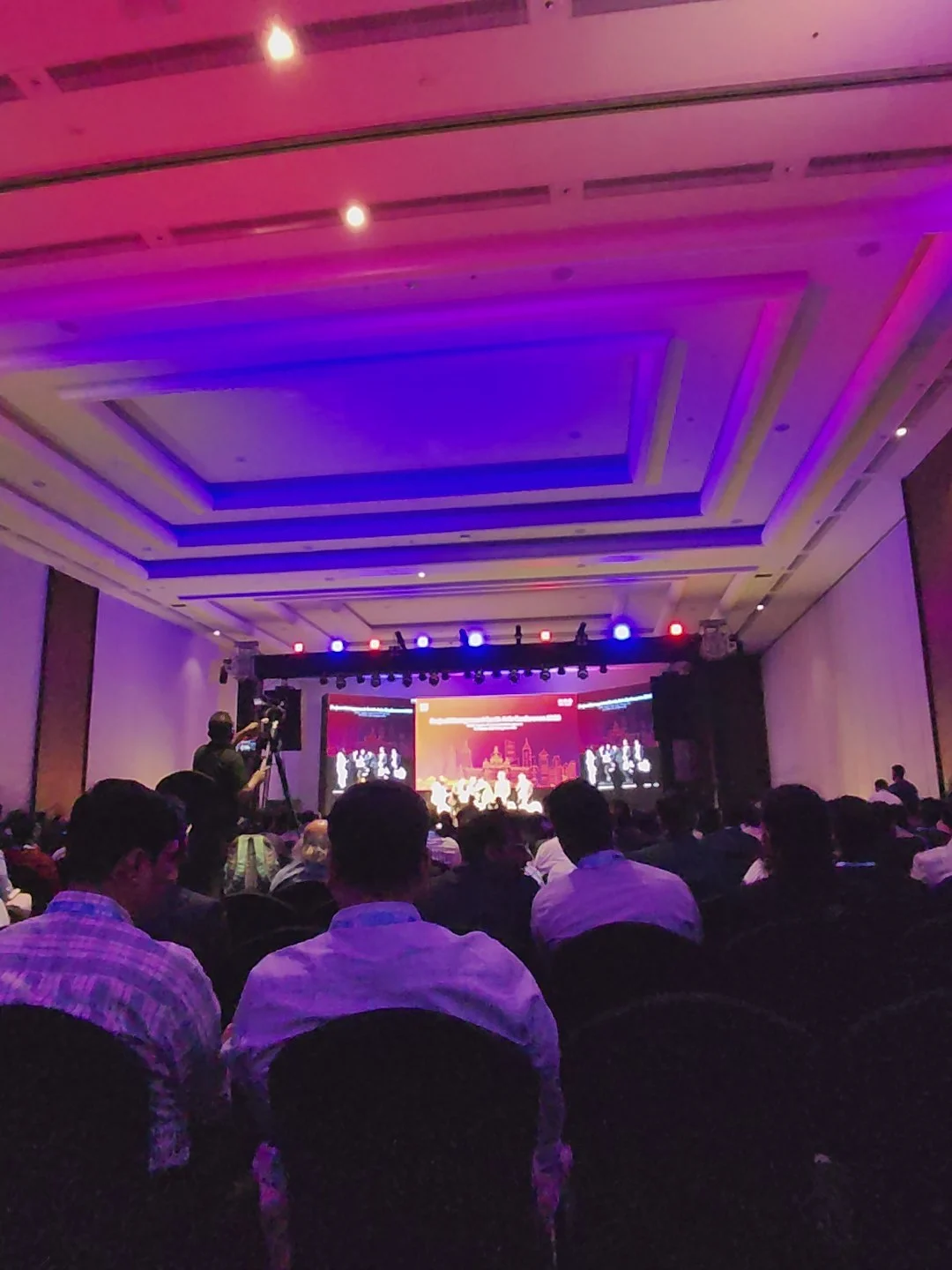 A large conference hall with an audience seated facing a brightly lit stage and screen, bathed in purple and blue lighting.