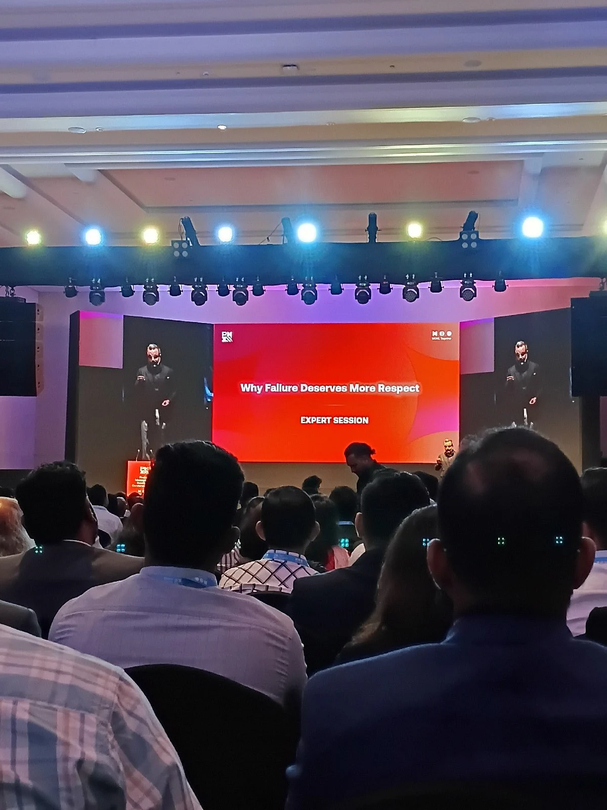 A conference audience watches a large red stage screen reading 'Why Failure Deserves More Respect' during an expert session.