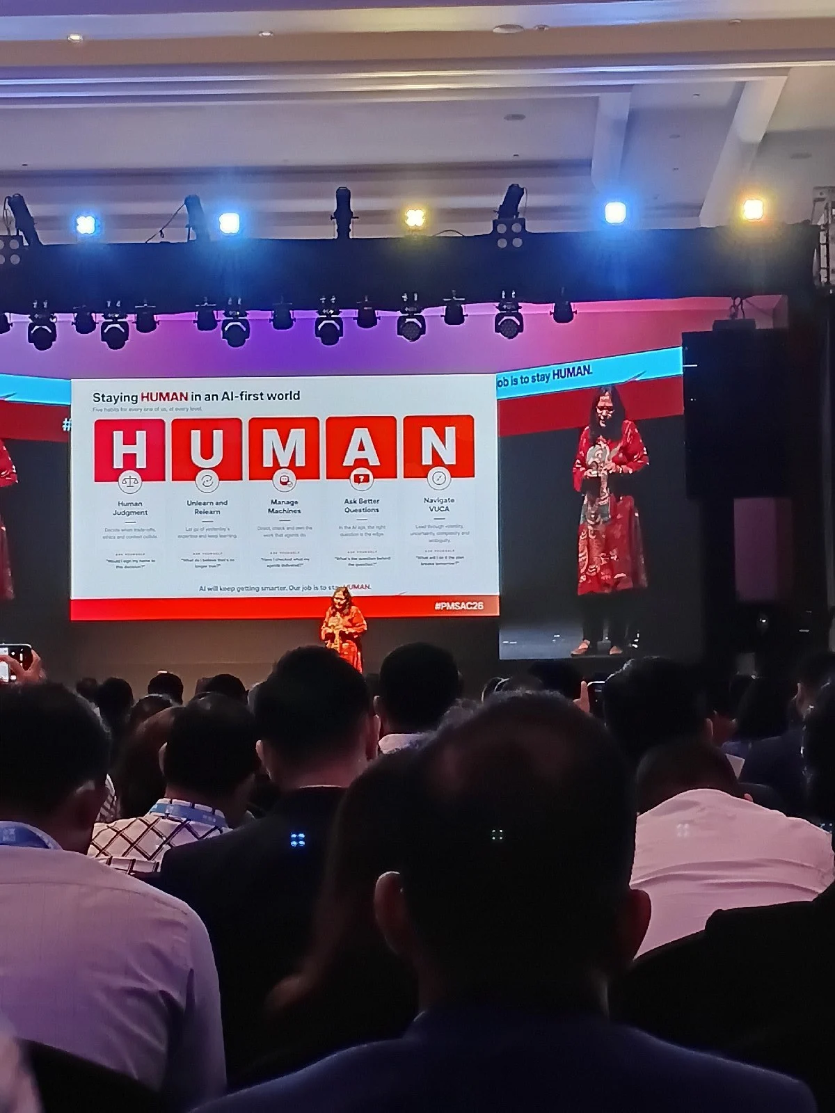 A speaker in a red outfit presents a slide titled 'Staying HUMAN in an AI-first world' on a large stage screen to a seated audience.