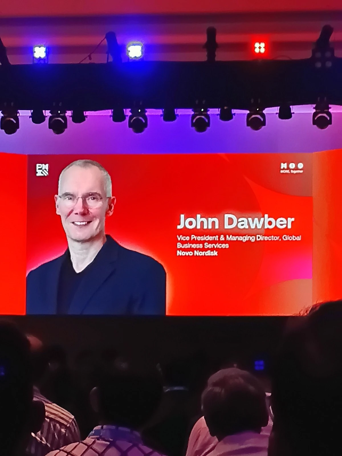 A large stage screen displays a speaker card with a portrait and name for John Dawber of Novo Nordisk, seen from the audience.