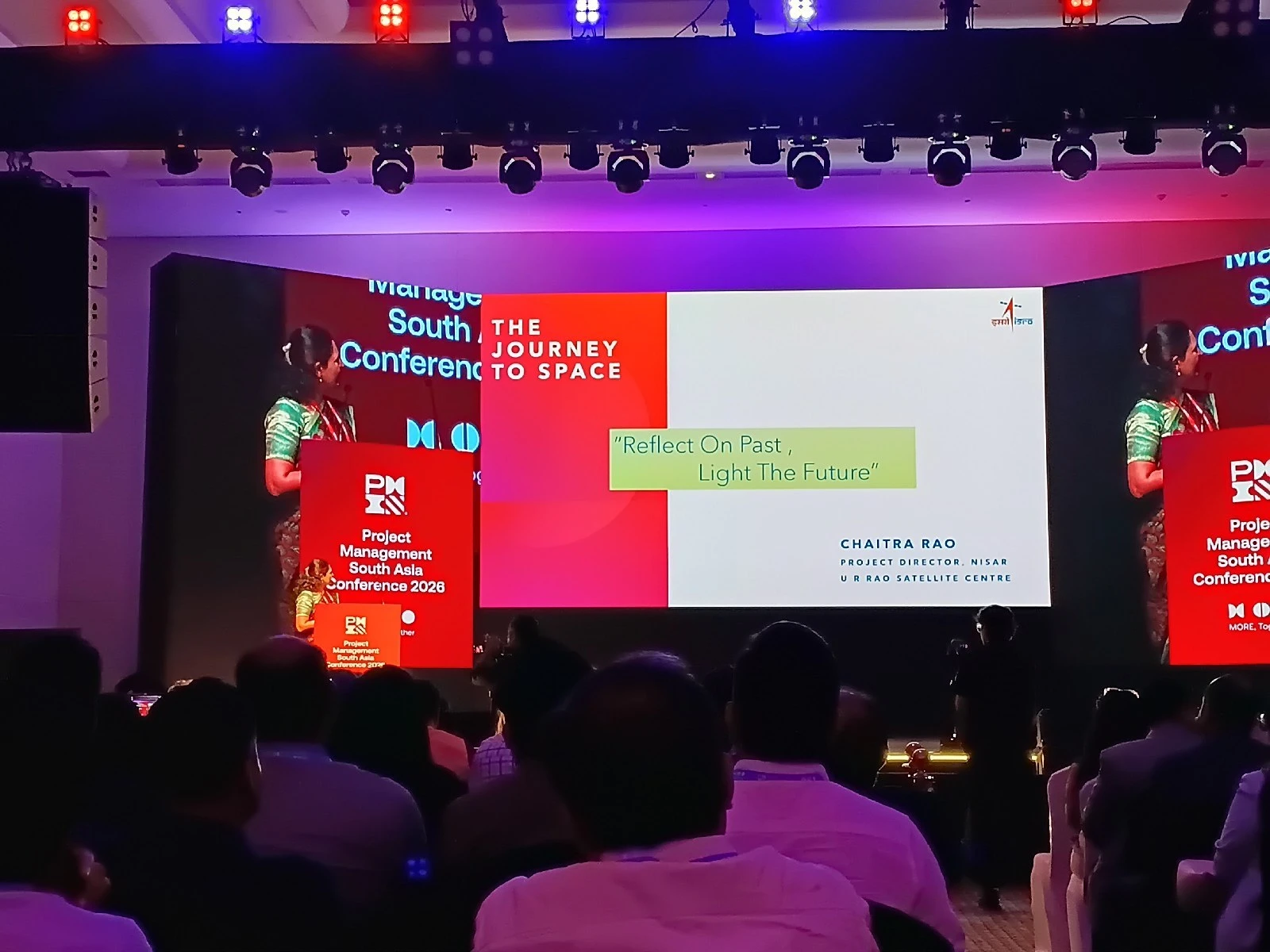 A conference audience watches a large stage screen displaying a speaker's title slide and a quote.