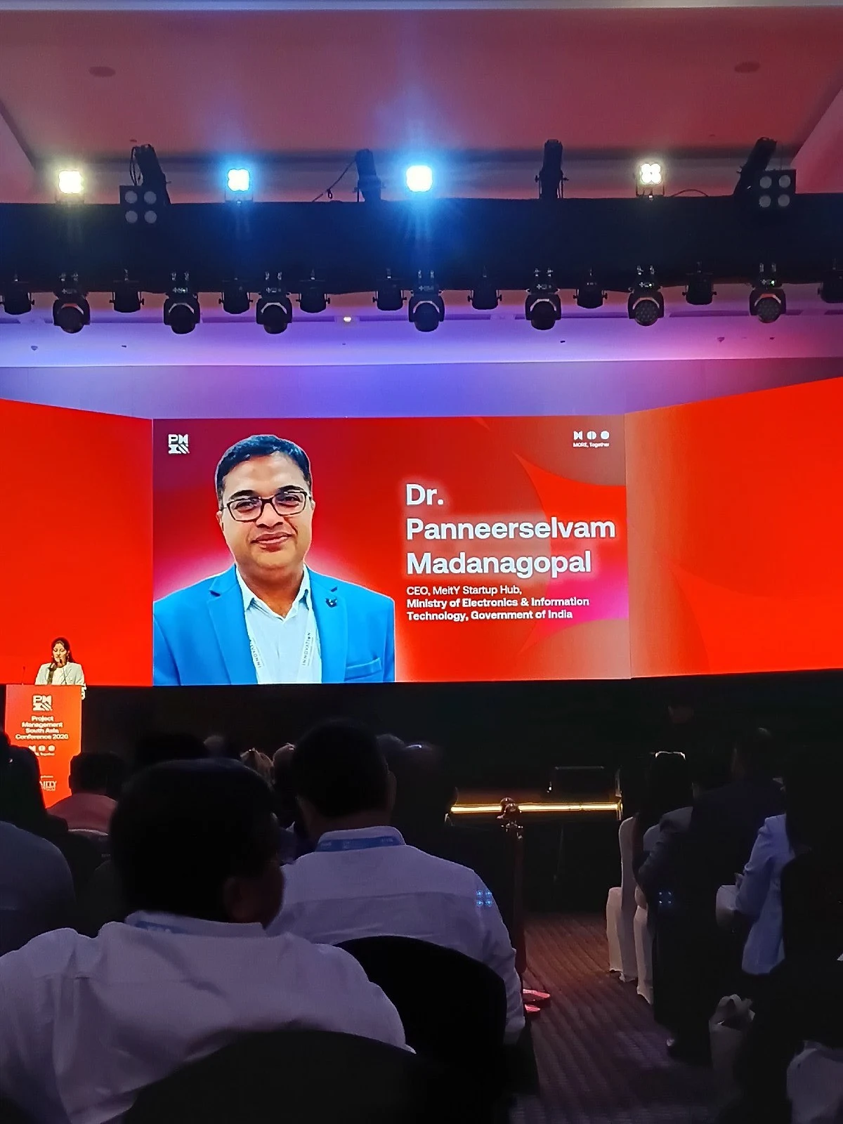 A large conference screen displays a speaker card for Dr. Panneerselvam Madanagopal while an audience watches and a person stands at a podium.