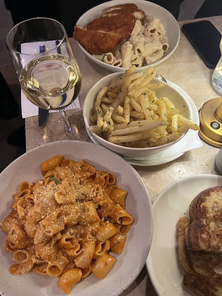 Table spread at a New York City restaurant featuring a bowl of rigatoni vodka pasta with breadcrumbs, a bowl of Italian fries with herbs, a glass of white wine, and a piece of toasted bread, shot from above.