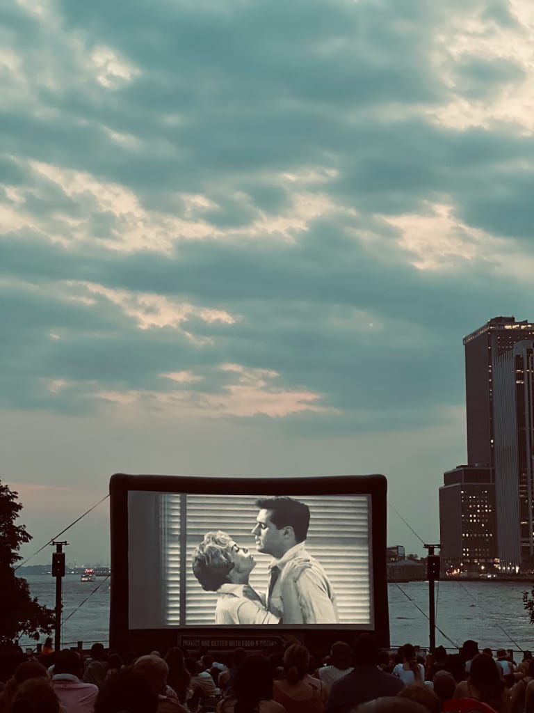 Outdoor movie screening in New York City showing a black and white classic film on a large inflatable screen, with the Manhattan skyline and East River visible in the background under a dramatic cloudy sky, with a crowd of people seated in the foreground.