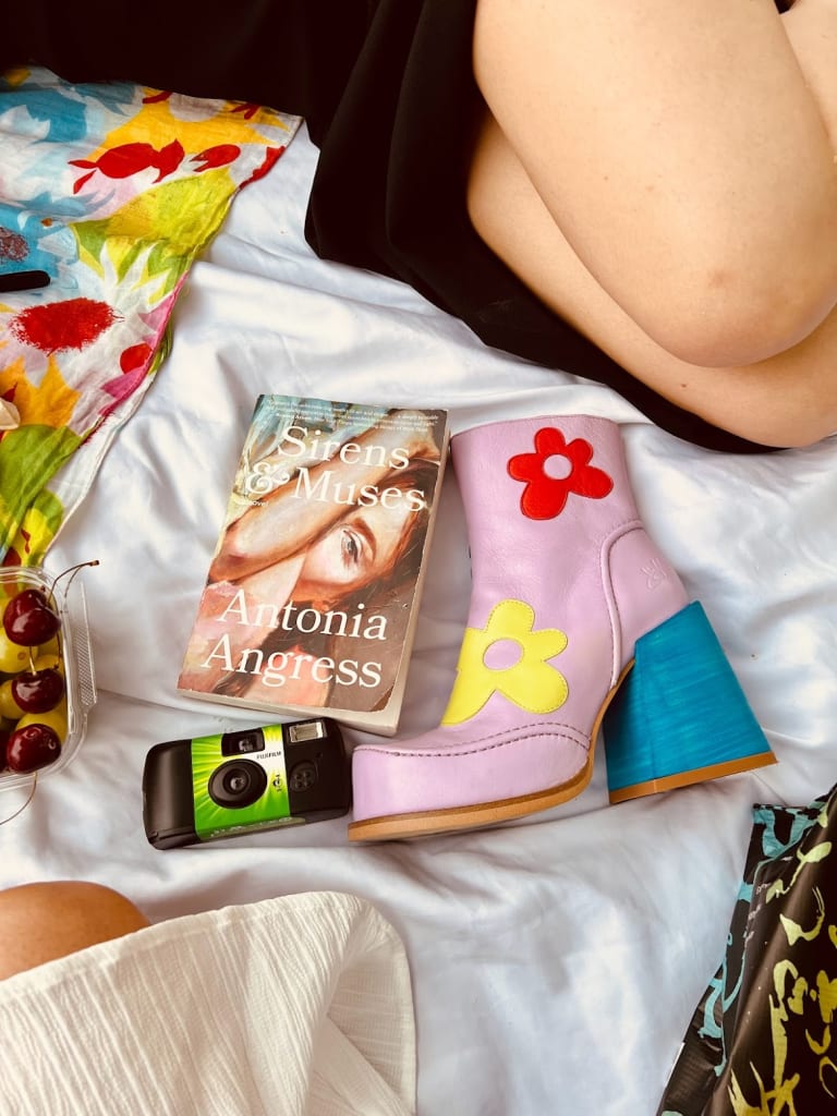 Flat lay photo on a white blanket in Central Park NYC featuring a copy of Sirens and Muses by Antonia Angress, a disposable film camera, fresh cherries, and a pair of lavender cowboy boots with floral embroidery, with a colourful floral scarf in the background.