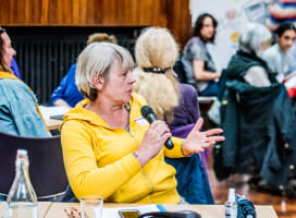 Person in yellow top talking into a microphone and gesturing in a busy room