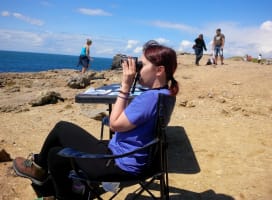 Young person sitting on a chair on a cliff, looking at the sea with binoculars
