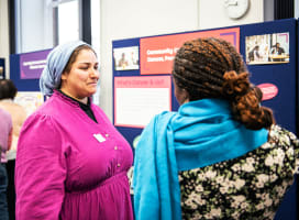 Two people in colourful clothing talking in front of a display board