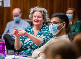 A female presenting person talking to someone off camera, someone is in the foreground listening whilst wearing a surgical mask