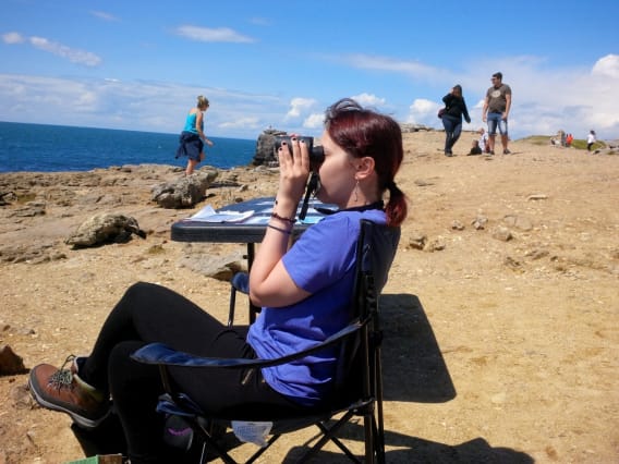 Young person sitting on a chair on a cliff, looking at the sea with binoculars