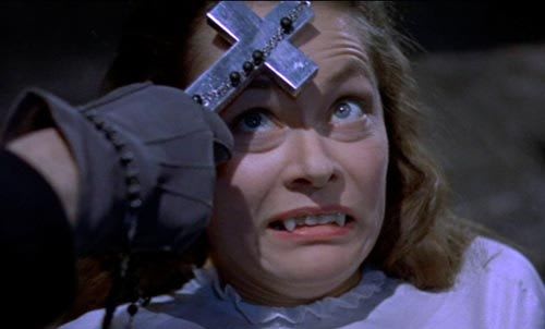 Scene from a horror movie showing a woman with a fearful, grimacing expression as someone holds a silver cross to her forehead. The image is from a classic exorcism or vampire film scene.