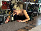 Anna working at a large machine, crafting a rug, with shelves of colourful yarn spools in the background.