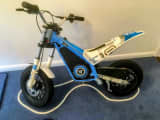 Blue and white electric motorbike parked on a blue carpet mat with a custom whipped edge, indoors.
