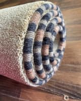A close-up of a rolled carpet with multi-coloured, striped whipped edging in black, tan, and brown, showing the texture and variety of yarns.