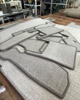 Stack of custom-shaped, grey helicopter mats with whipped edging, displayed in a workshop with carpet rolls in the background.