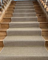 Staircase with a custom-fitted, neutral-toned runner featuring whipped edges and brass stair rods, installed on wooden steps.
