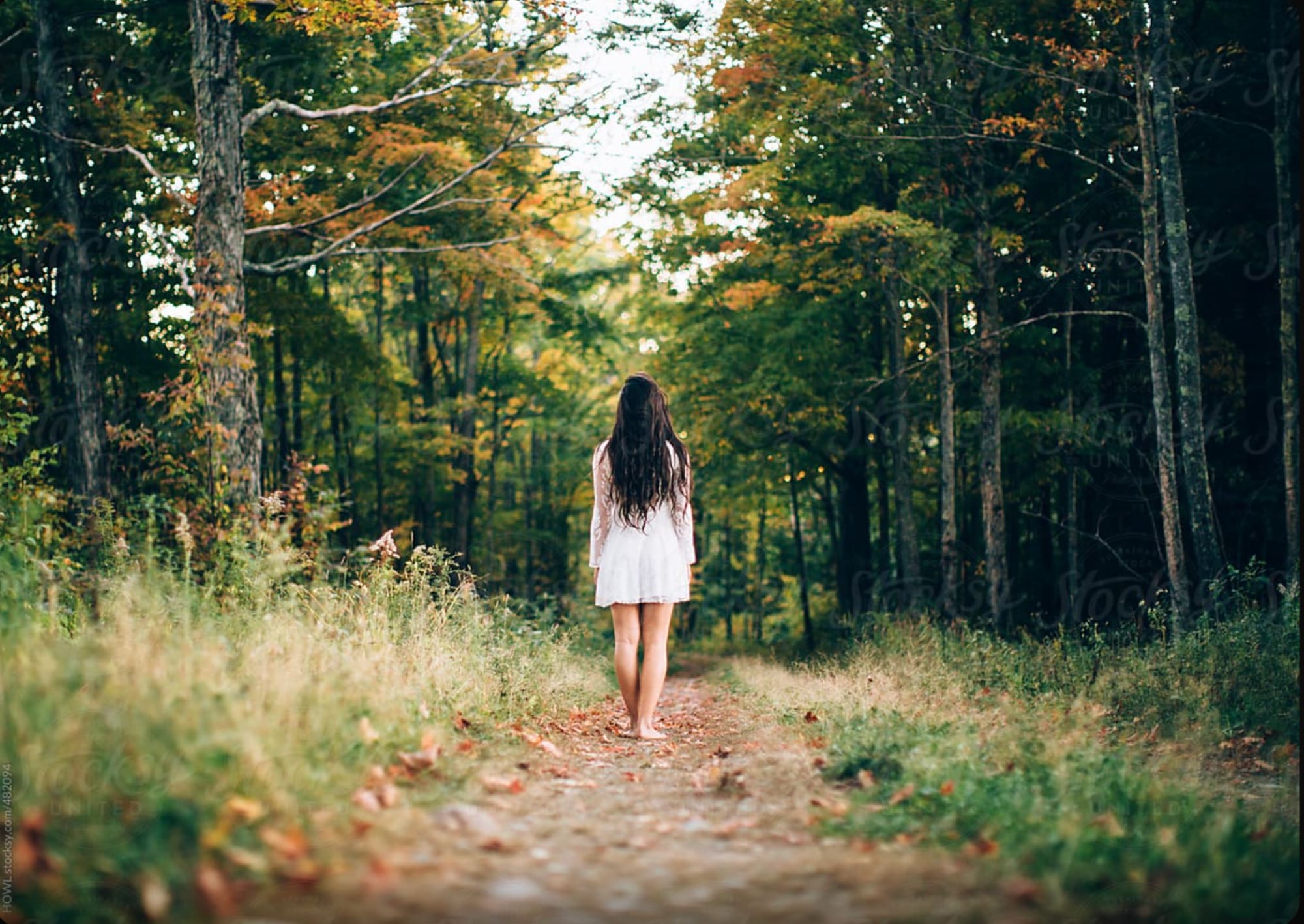A woman in a white dress walking alone down an autumn forest path, soft light filtering through the trees