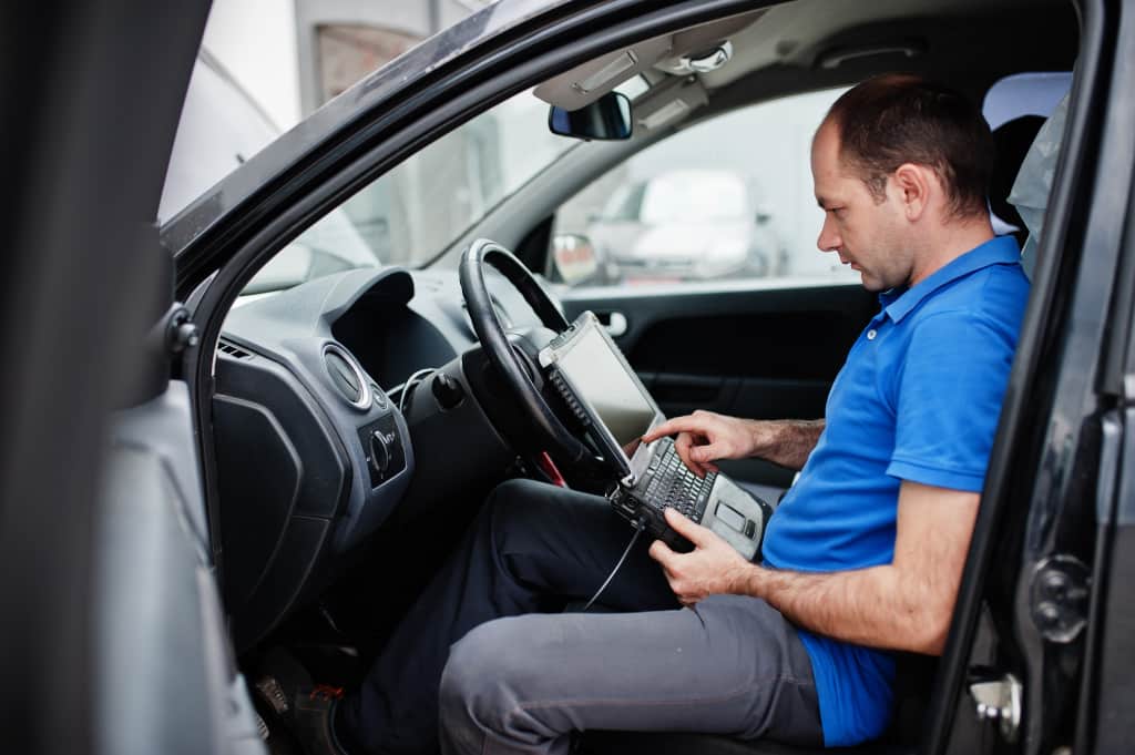Car repair and maintenance theme. Electric mechanic in uniform working in auto service, making car diagnostics using obd device with laptop.