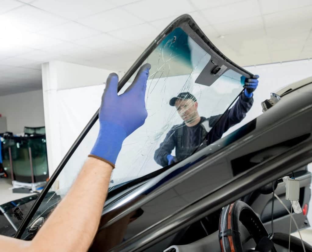 Two technicians installing a new windshield on a car inside a brightly lit auto repair shop, wearing gloves for safety and precision.