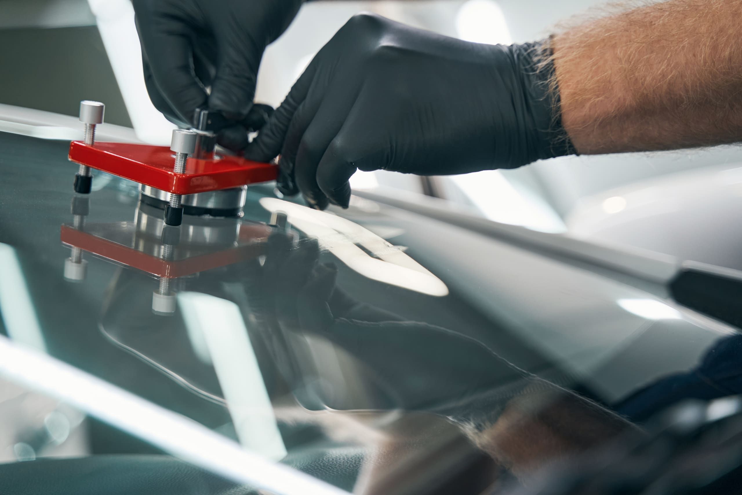 Close-up of a technician using a windshield repair kit to fix a chip in a car's windshield, wearing black gloves for safety in a Michigan auto repair shop.